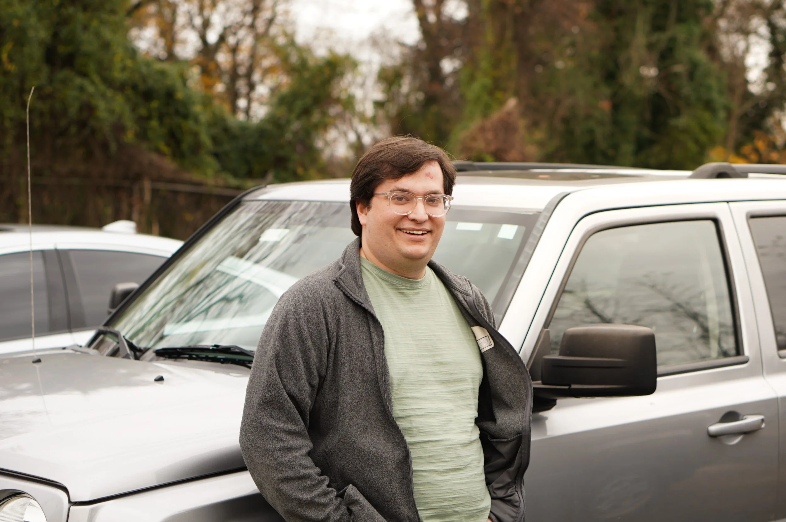 A man with glasses smiling and standing in front of a silver SUV during daytime, with autumn trees in the background.