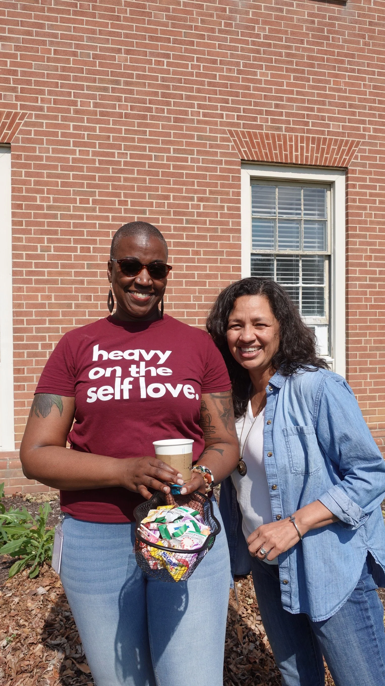 Two women standing outdoors in front of a brick building smiling at the camera. One woman with short hair, wearing sunglasses and a maroon t-shirt with white text, holding a coffee cup and a basket of snacks. The other woman with curly hair, wearing a blue denim shirt over a white top.