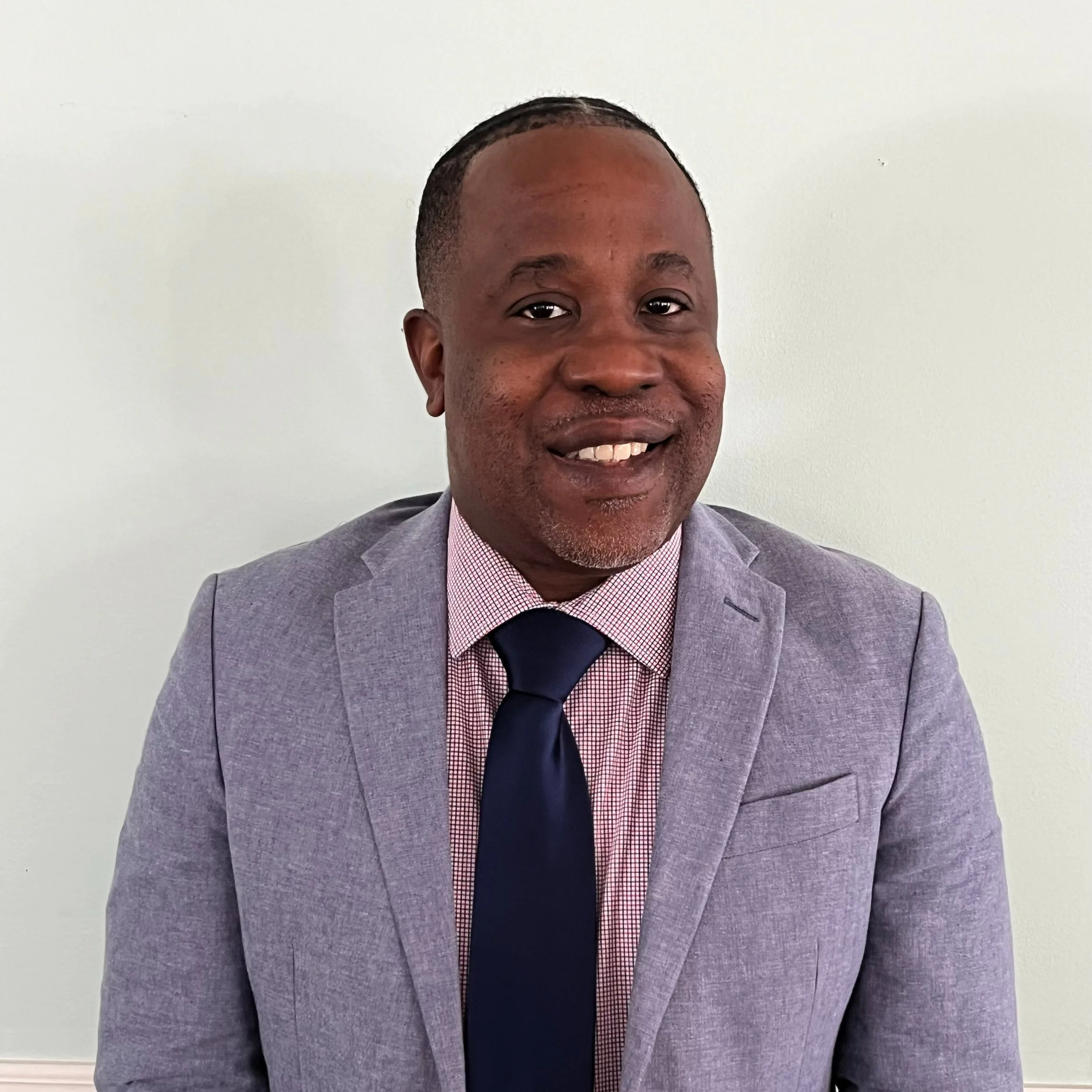 A man in a gray suit jacket, pink checkered shirt, and dark tie smiling at the camera, standing against a plain white wall.