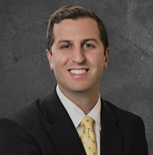 A smiling man in a suit with a white shirt and yellow tie posing against a gray wall.