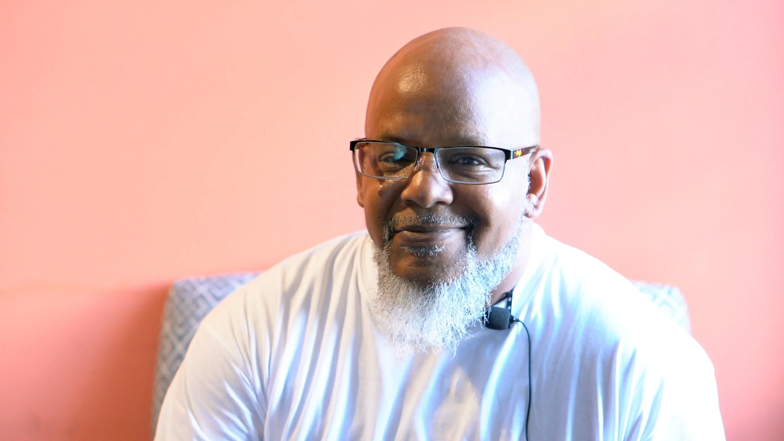 A middle-aged man with a bald head, glasses, and a gray beard smiling, wearing a white shirt with a headset microphone, sitting against a pink background.
