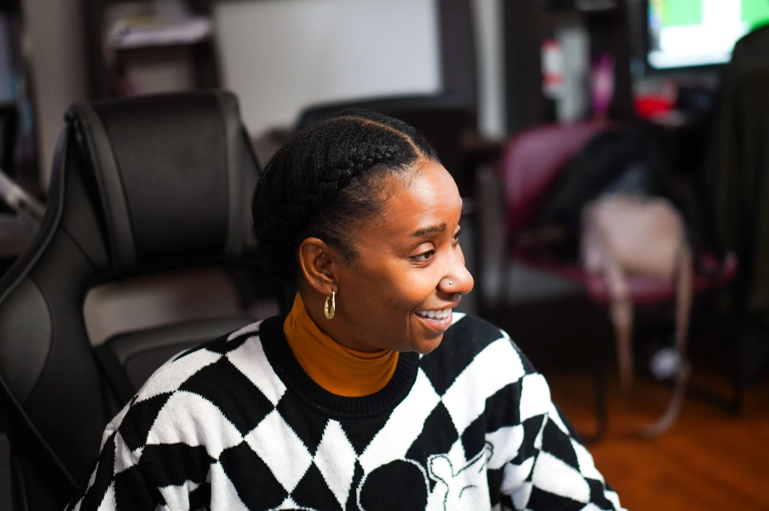 A woman with dark skin and braided hair is smiling while sitting in a black chair in an indoor setting.