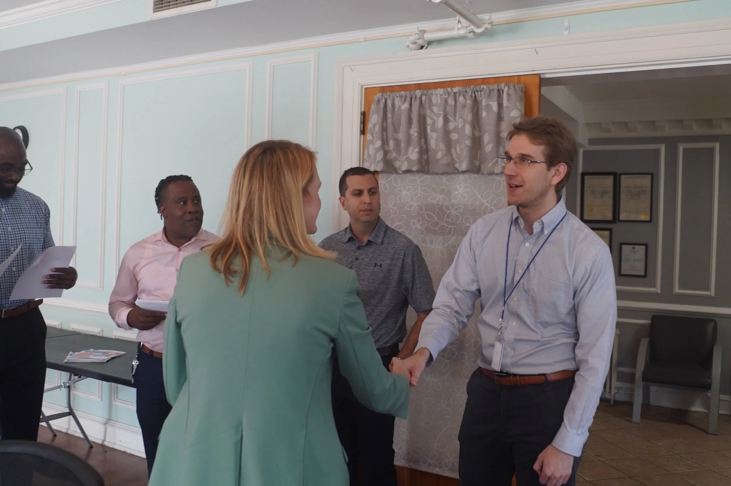 A group of people interacting in a professional setting, including a woman and a man shaking hands and others holding papers, in a room with framed certificates on the wall.