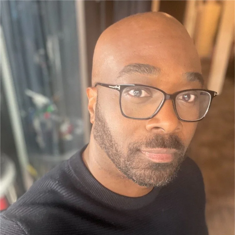 A close-up selfie of a man with a shaved head, glasses, and a beard, wearing a black shirt in an indoor setting.