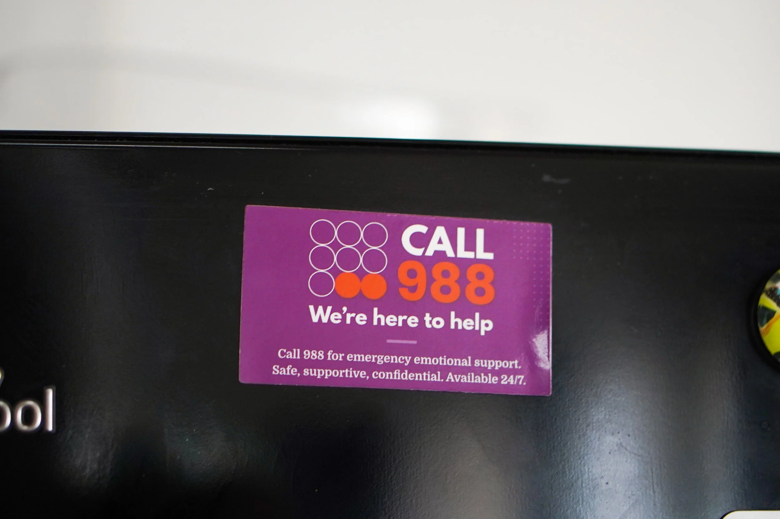 Close-up of a purple sticker on a black surface with text that reads, 'CALL 988 We're here to help. Call 988 for emergency emotional support. Safe, supportive, confidential. Available 24/7.'