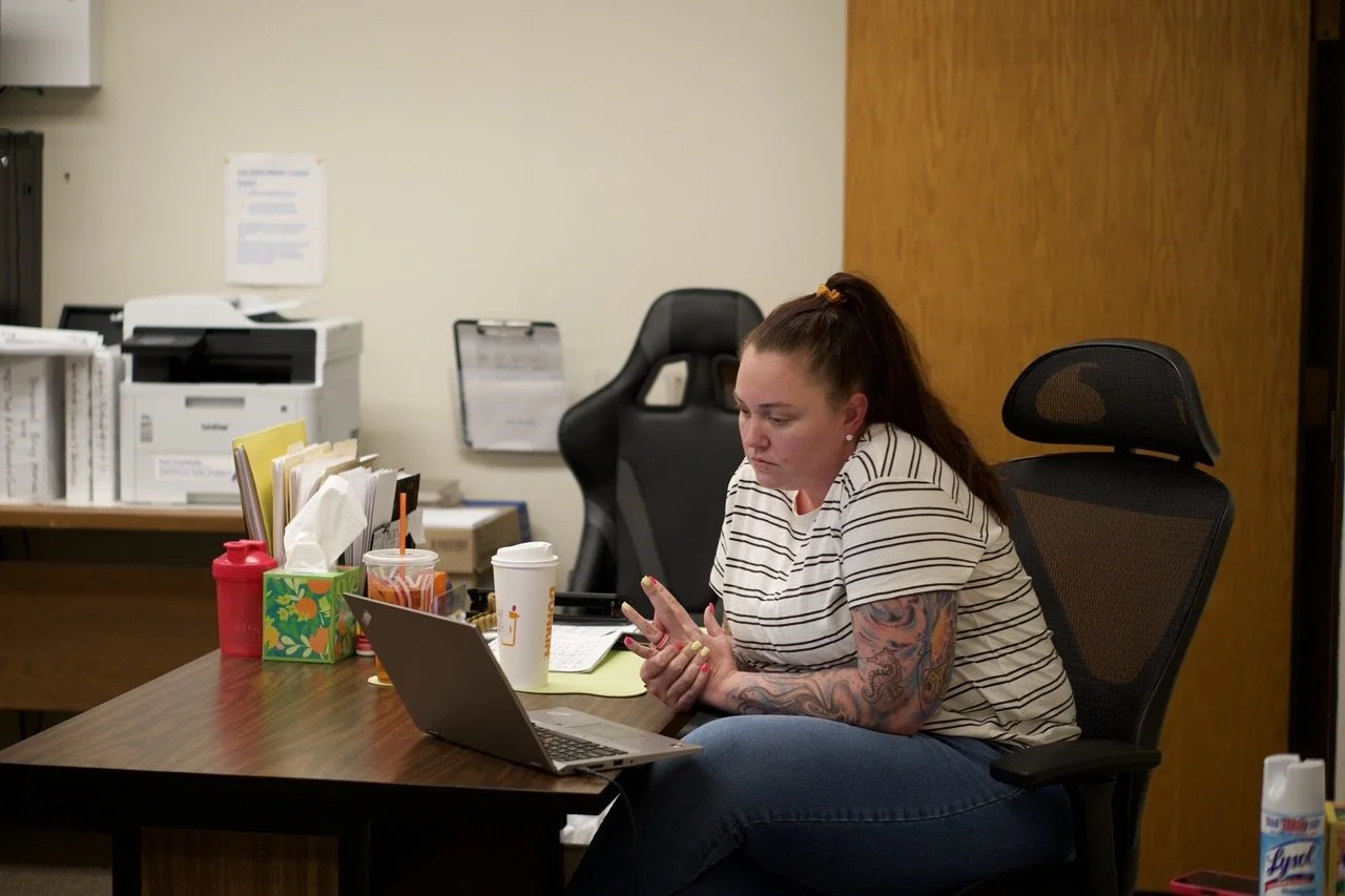 A woman with long dark hair in a high ponytail, wearing a striped shirt and jeans, is sitting at a desk with a laptop in an office. She has tattoos on her arms and is looking at her phone. The desk has various items including a red water bottle, tissue box, cups, and papers. There are office equipment and filed papers in the background.