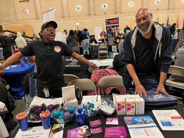 Two men standing behind a table at an indoor event, smiling and talking. The table displays health-related products, pamphlets, and promotional materials. People and booths are visible in the background.