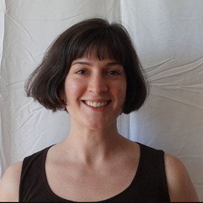 A woman with short brown hair smiling, wearing a black sleeveless top, standing in front of a white backdrop.