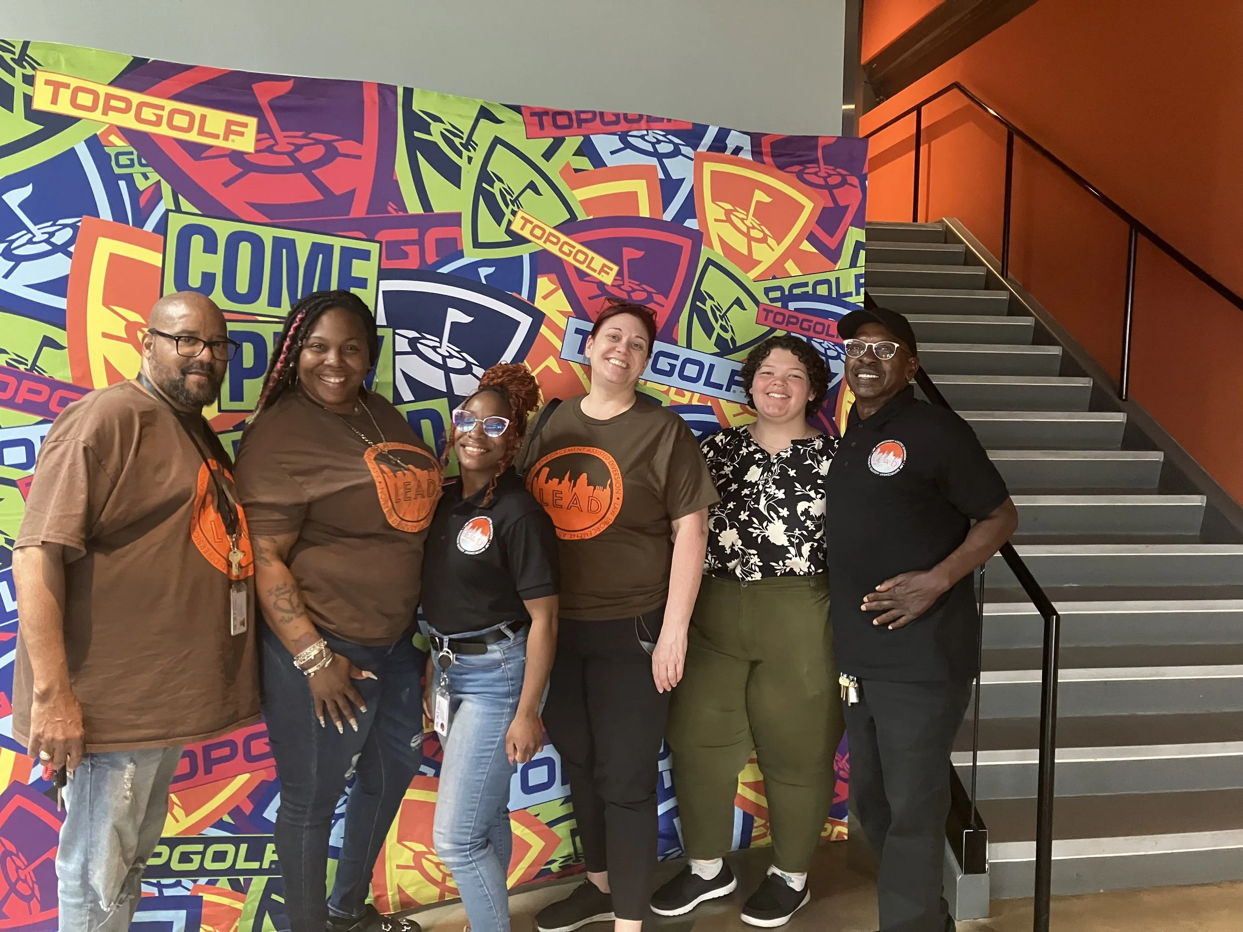 A group of six diverse people standing together in front of a colorful backdrop with references to golf and the words "COME GOLF" and "TOP GOLF" on it, smiling and posing for the photo.