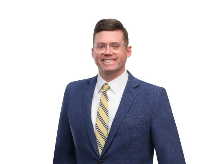 A man in a blue suit and yellow striped tie smiling, wearing glasses, against a white background.