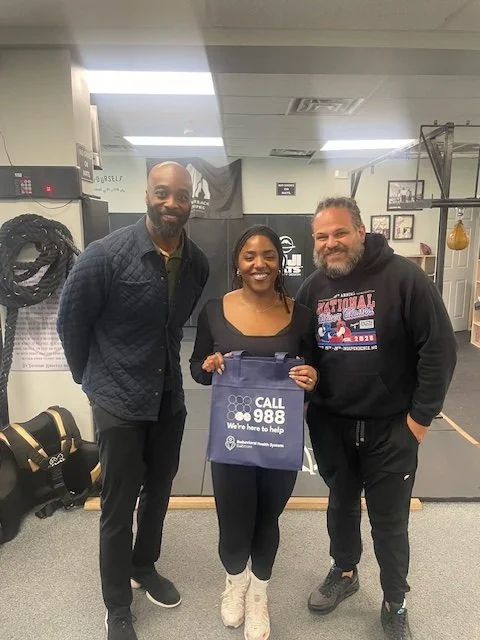 Three people smiling and standing inside a gym, with one woman holding a blue bag that reads 'Call 988 We're here to help'.