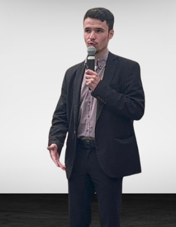 Homme debout parlant en microphone lors d'une présentation. Il porte un costume et une chemise.