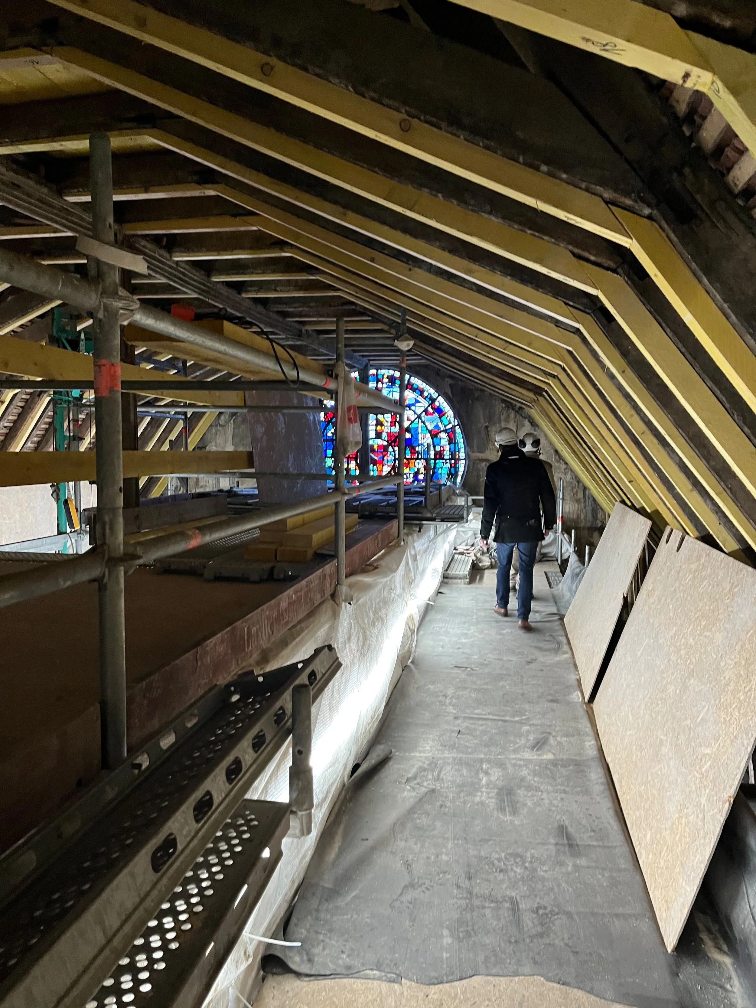 À l'intérieur d'une ancienne église en rénovation, un homme marche dans le passage entre des échafaudages et des panneaux de contreplaqué. Derrière lui, une grande rosace colorée en vitrail.