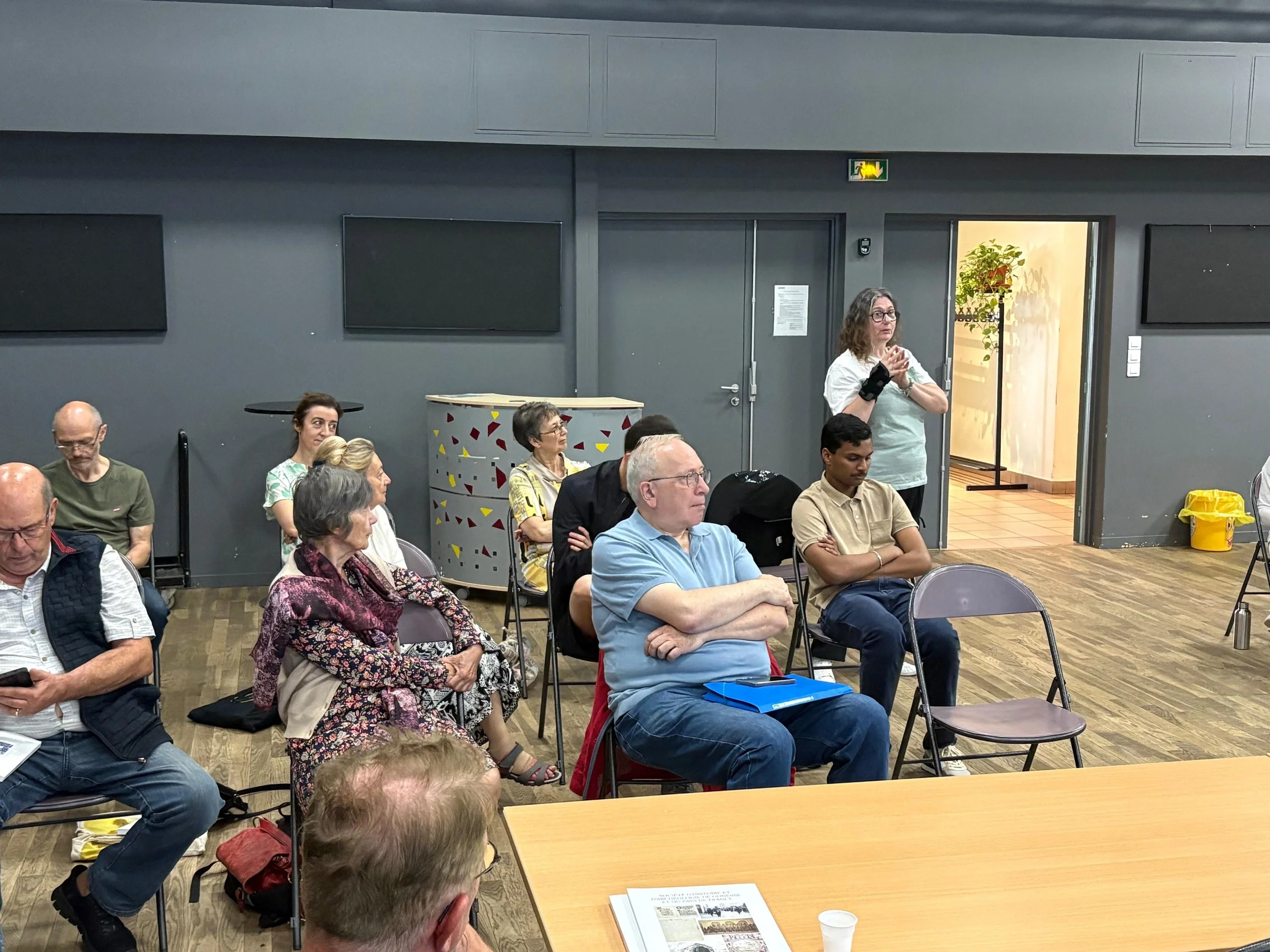 Une salle de réunion avec plusieurs personnes assises en train d'écouter une présentation. Une femme se tient debout à côté d'une porte, notamment une plante visible dans la pièce derrière elle. La salle a des murs gris et une ambiance de conférence.
