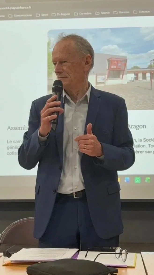Un homme en costume parlant dans un micro lors d'une présentation ou conférence avec un fond montrant un diaporama