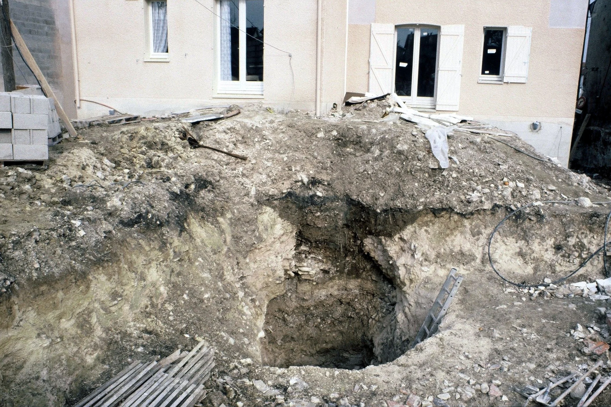Une excavation profonde dans le sol devant une maison en construction ou en rénovation, avec des tuyaux en acier visibles au fond de la tranchée.