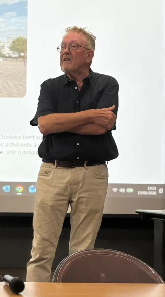 Un homme âgé avec des lunettes, portant une chemise noire et un pantalon beige, se tient avec les bras croisés dans une salle de présentation.