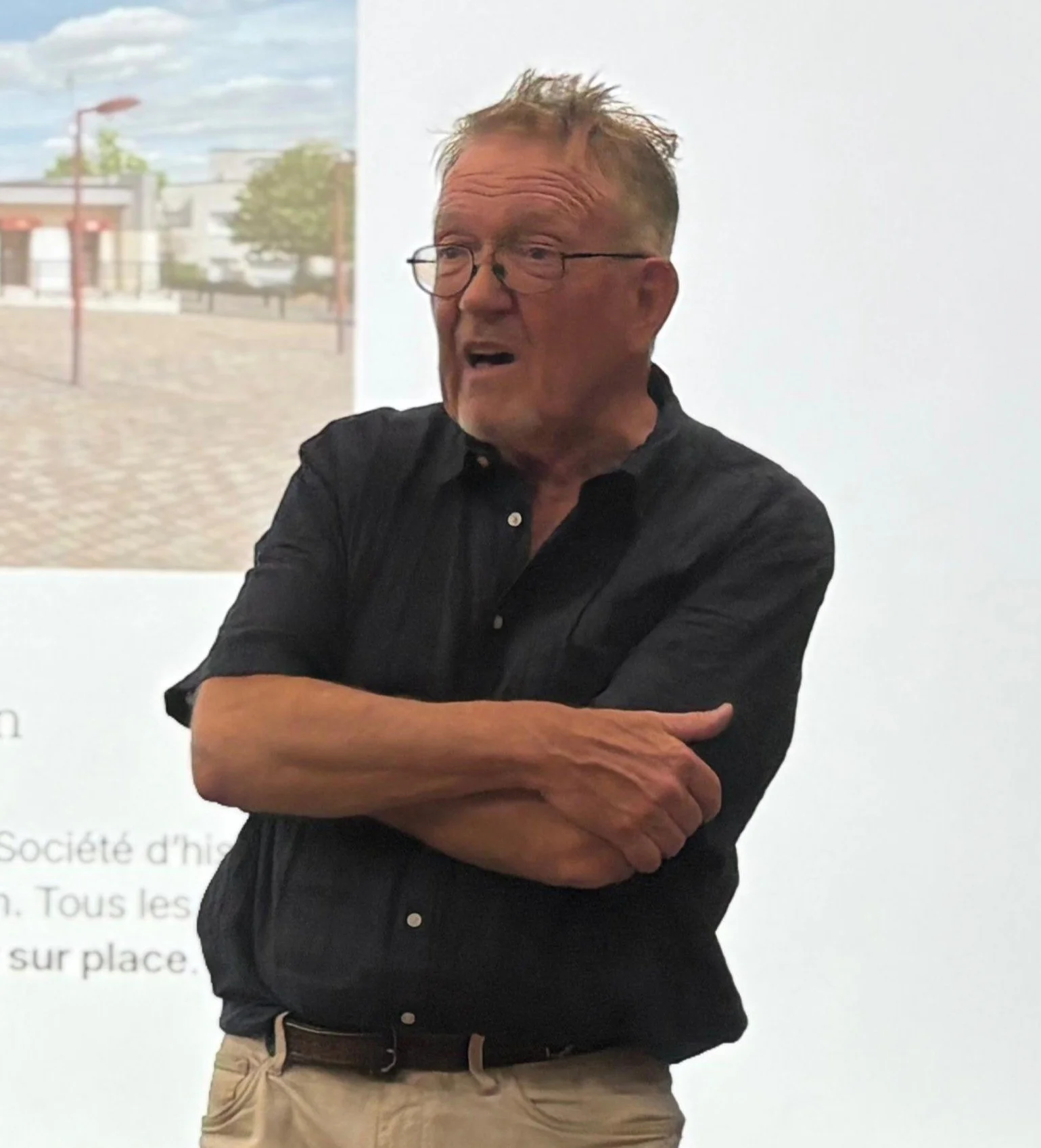 Un homme âgé avec des lunettes, portant une chemise noire et des pantalons beige, parle avec une expression animée, bras croisés devant une présentation ou un écran.
