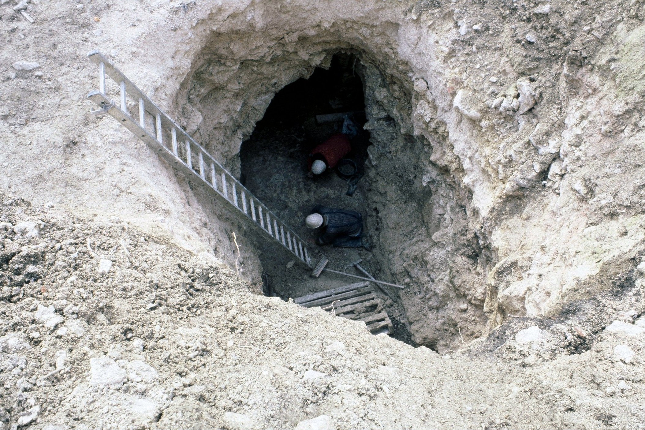 Une excavation avec deux personnes travaillant à l'intérieur, une échelle en bois et un casque de sécurité visible.