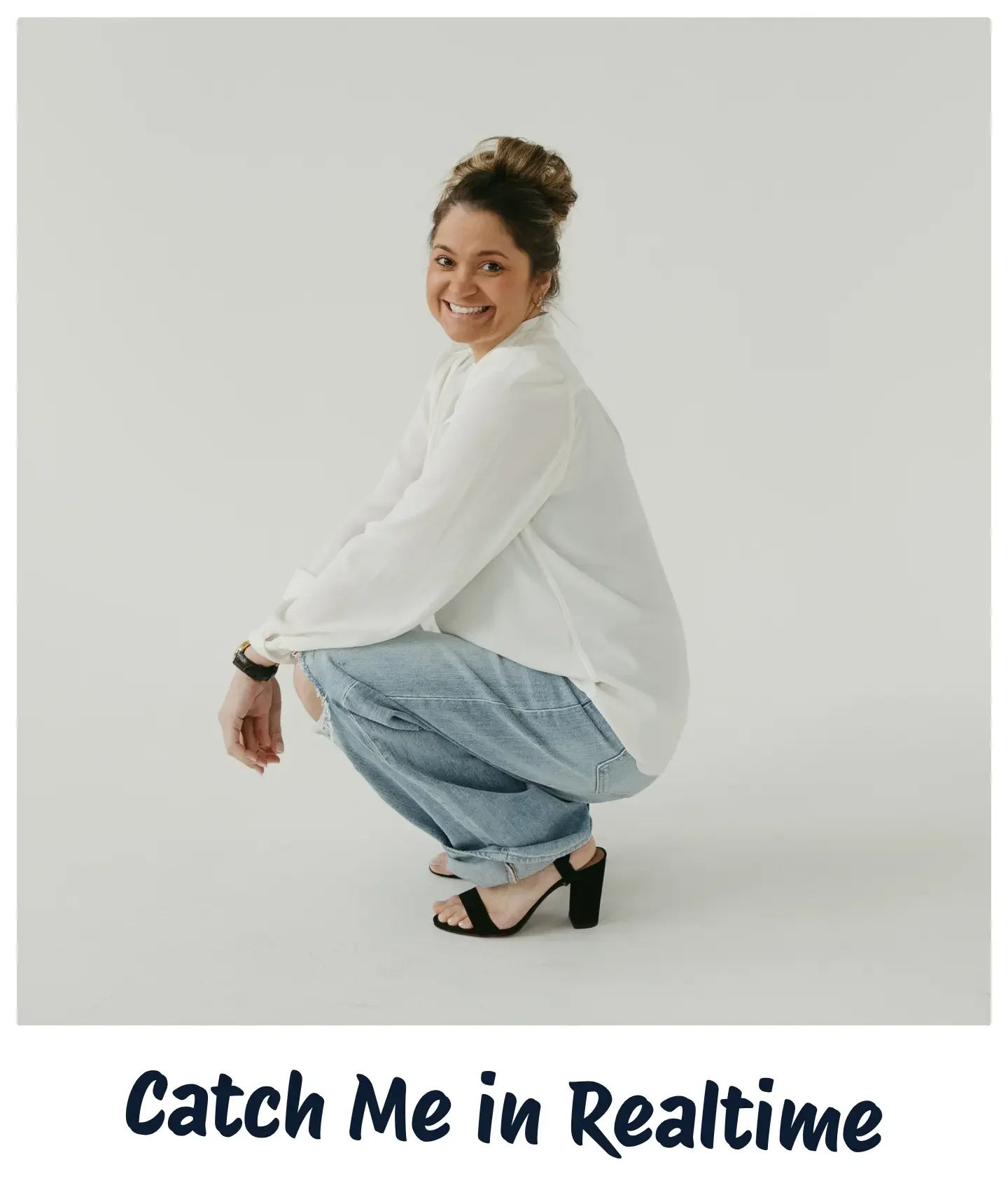 A woman with dark hair in a messy bun, smiling, squatting in a white shirt, ripped jeans, and black high-heeled sandals against a plain white background. Text below reads 'Catch Me in RealTime'.