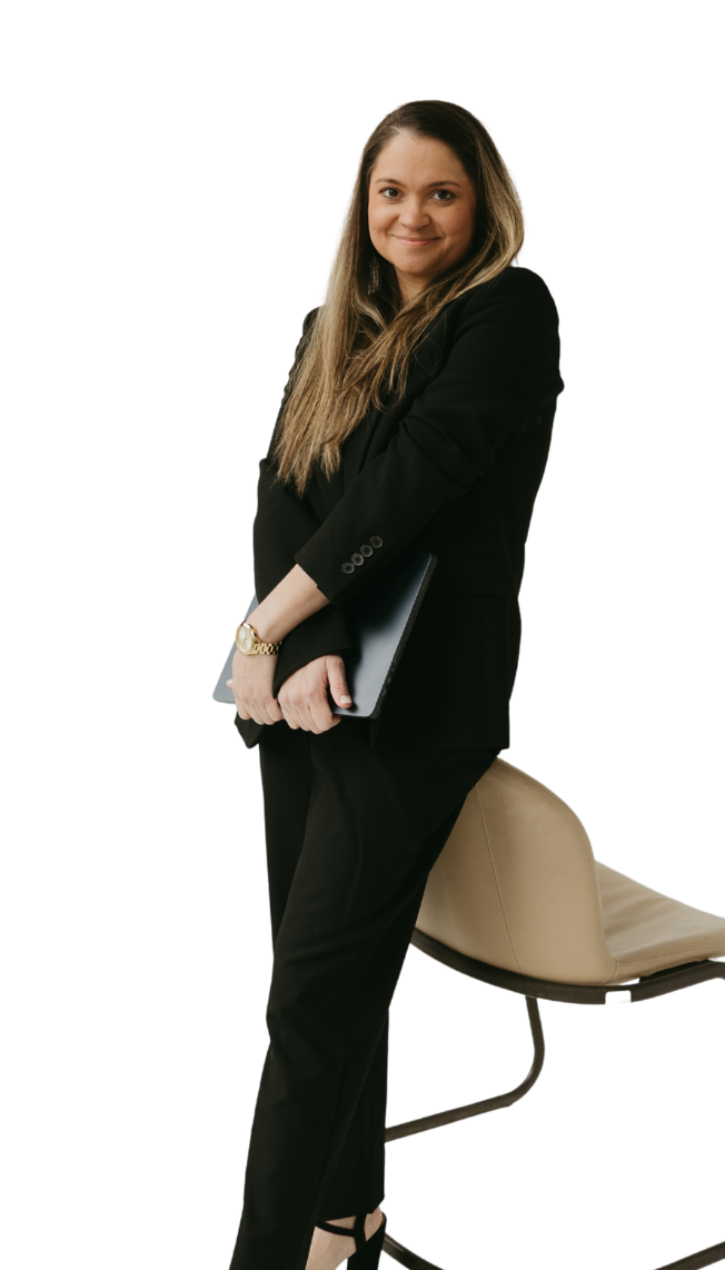 A woman with long, light brown hair in business attire standing next to a beige chair, holding a laptop, and smiling at the camera.
