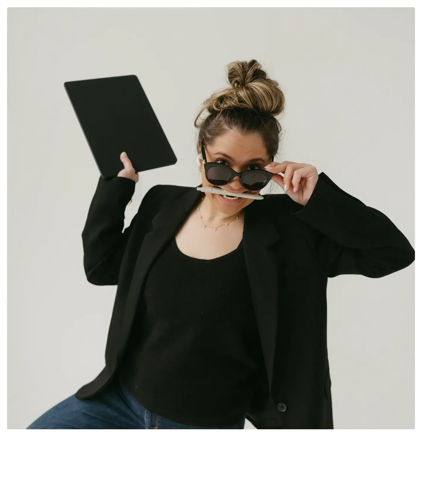 Woman with a top knot hairstyle, wearing sunglasses, a black blazer, black top, jeans, and a necklace, holding a tablet and smiling.