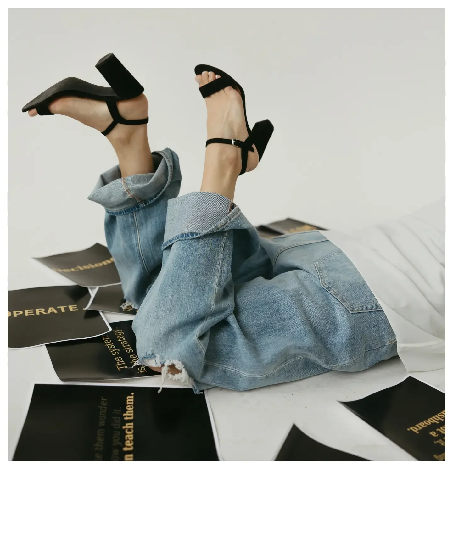Person lying on a white surface with legs raised, wearing ripped jeans and black high-heeled sandals, surrounded by black posters with gold text.