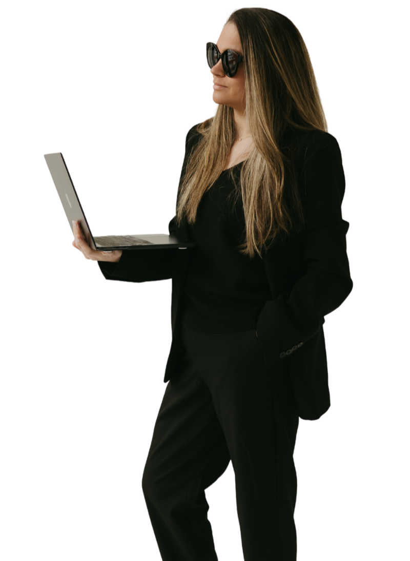 A woman with long, wavy hair, wearing large sunglasses and a black suit, holds a laptop in her right hand while standing against a plain background.