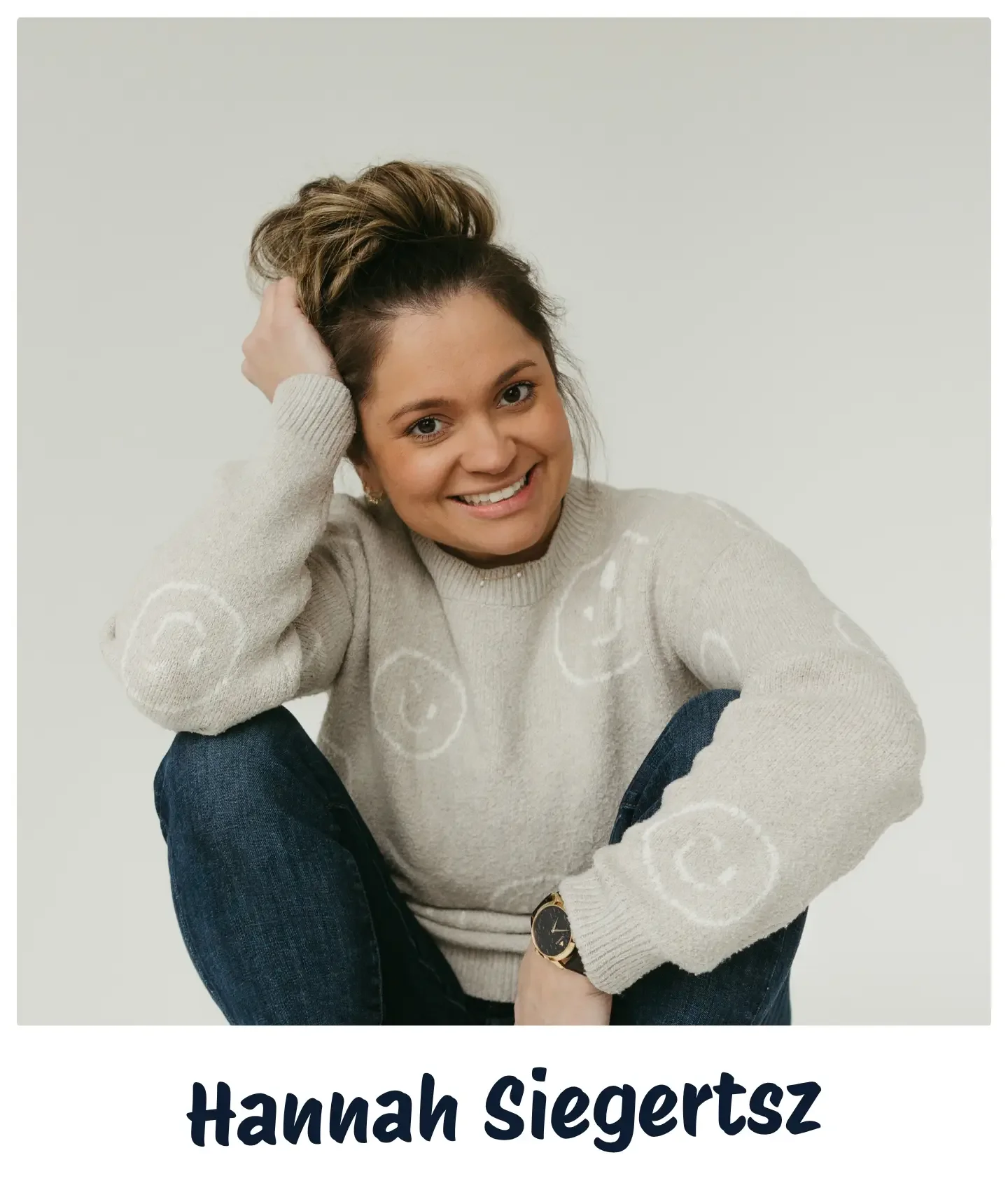 A woman with brown hair in a messy bun, smiling and sitting in a casual pose against a plain background. She is wearing a beige sweater with swirl designs and blue jeans, with a watch on her left wrist. Her name, Hannah Siegertsz, is written in bold black text at the bottom of the image.