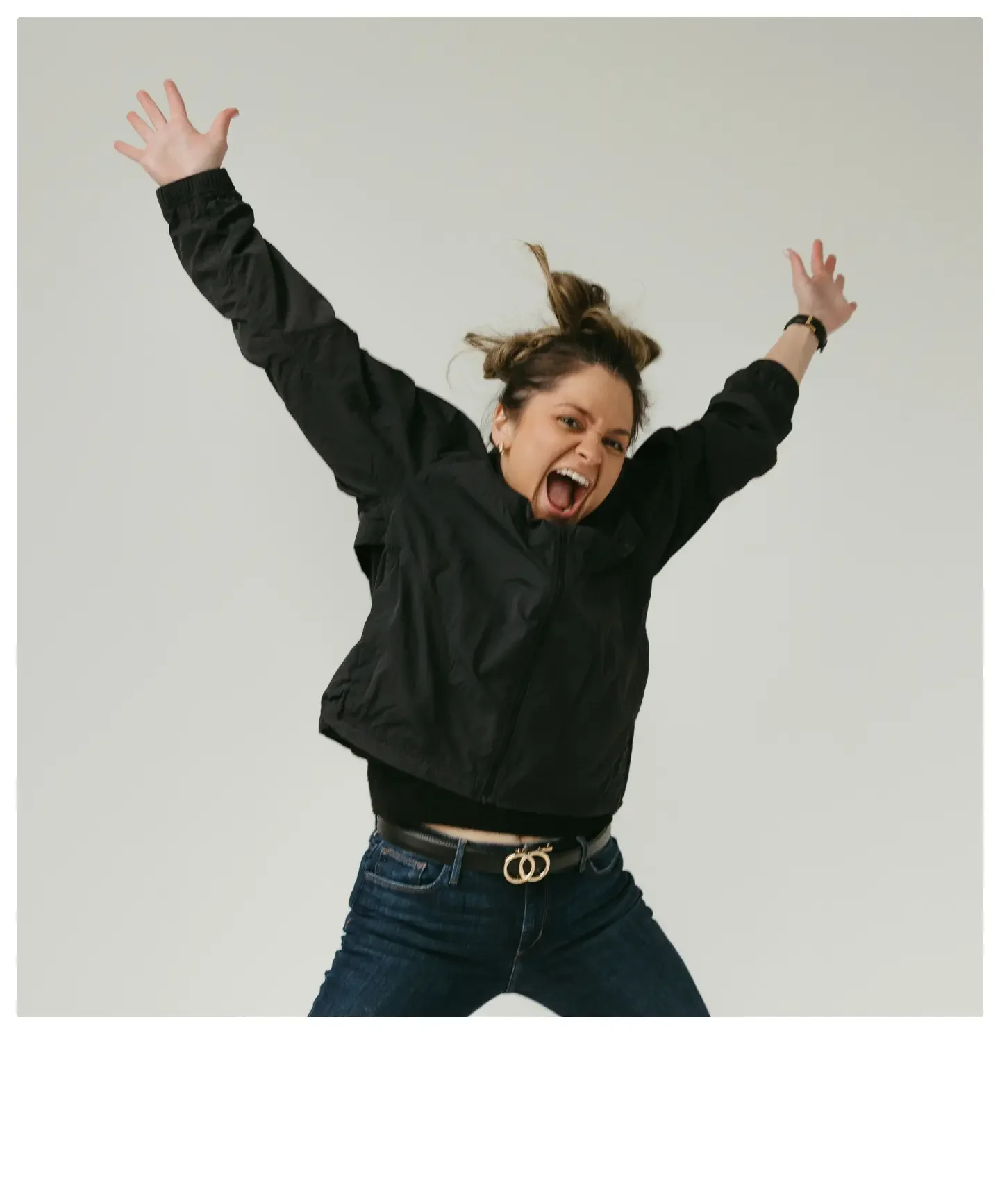A woman wearing a black jacket and dark jeans is jumping with her arms raised and an excited expression on her face against a plain white background.