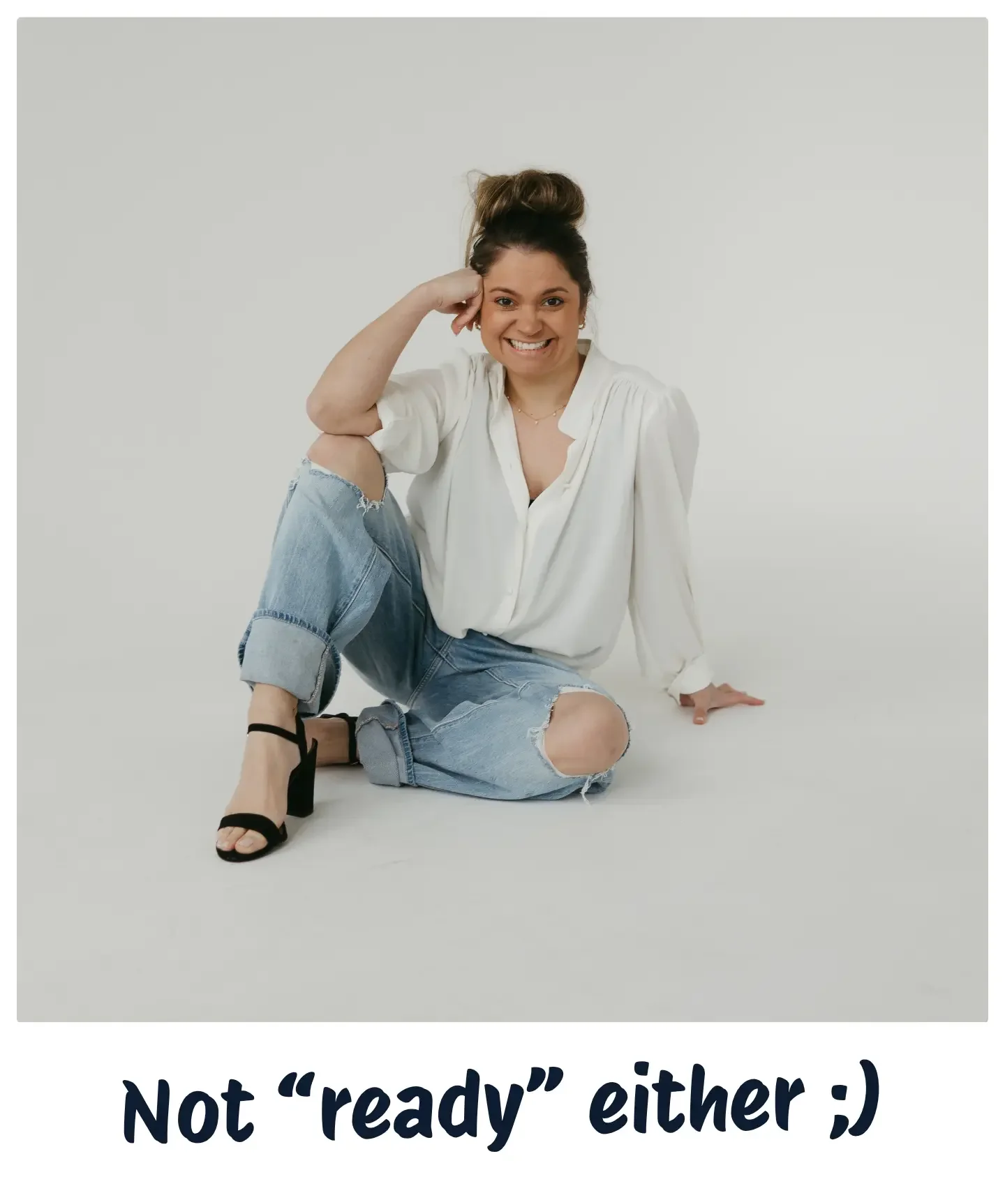 A woman sitting on the floor, smiling, wearing a white blouse, ripped jeans, and black high-heeled sandals, with her hair in a bun and a plain white background.