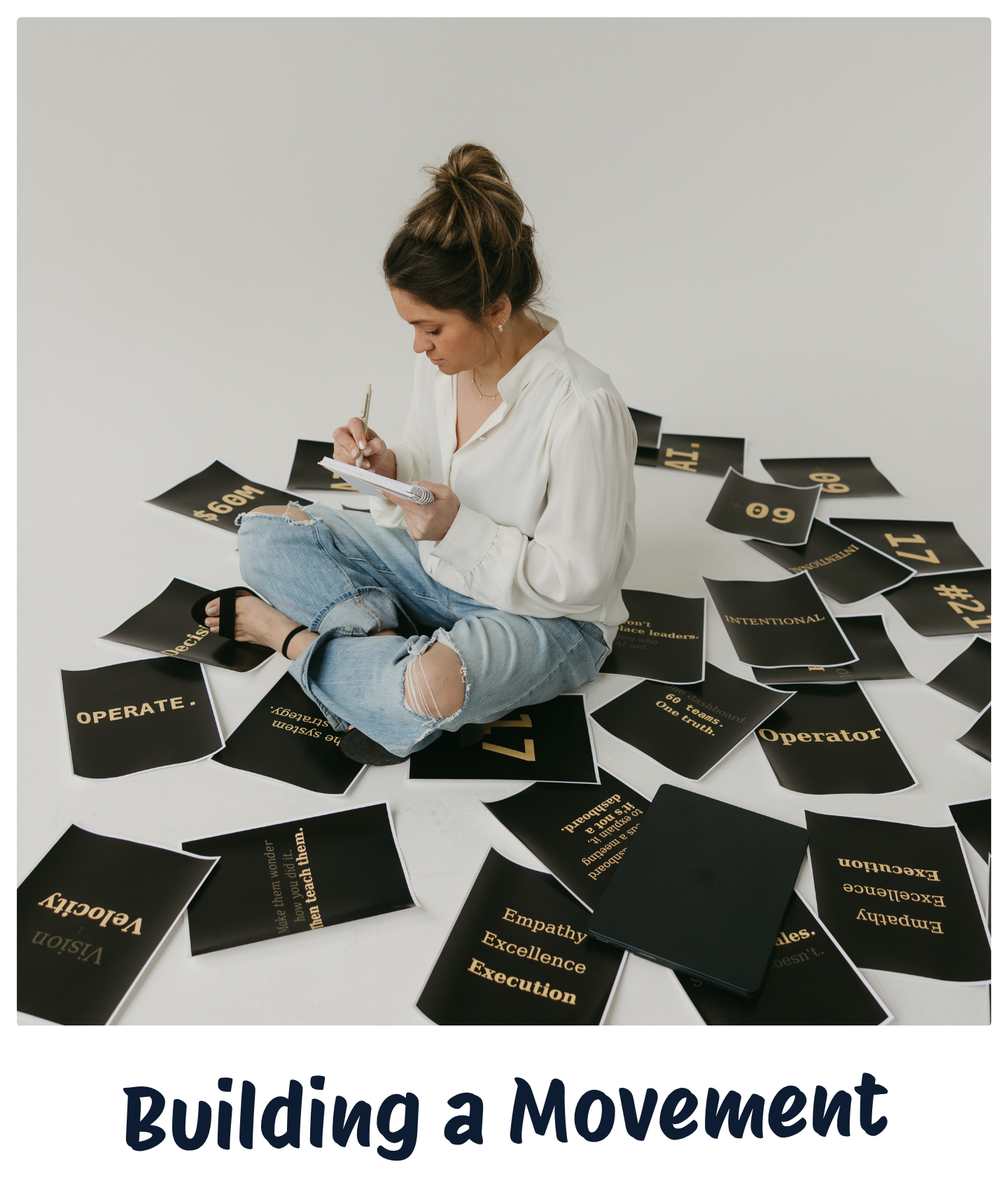 A woman sitting cross-legged on the floor, surrounded by black cards with gold text, writing in a notepad.