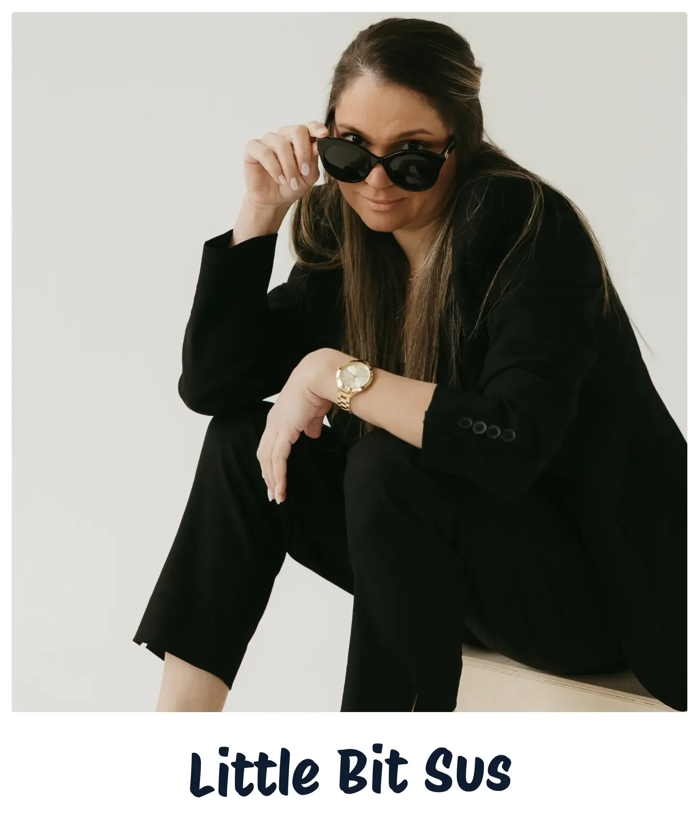 A woman with long brown hair, wearing black sunglasses, a black blazer, and black pants, sitting with one knee up, looking at the camera and adjusting her sunglasses. She is also wearing a gold watch on her wrist. The background is plain and light-colored. The text at the bottom reads "Little Bit SUS".