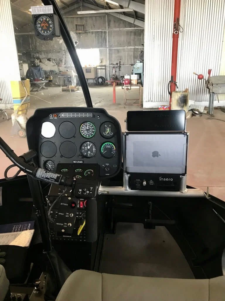 Cockpit of a helicopter with various gauges, controls, and screens, including an iPad mounted on the right side in an industrial hangar.