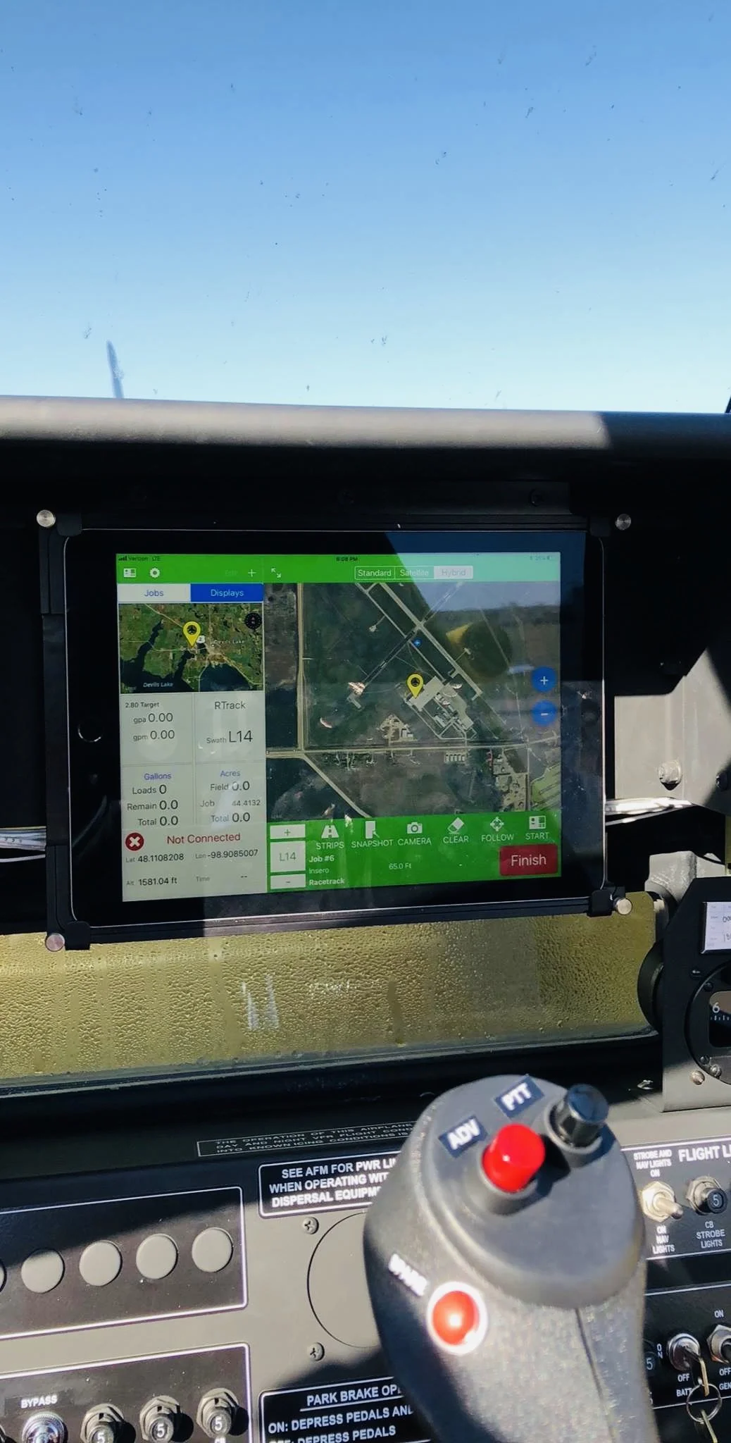 Dashboard screen inside a vehicle showing a map with a yellow marker near a racetrack, various controls, and a bright blue sky outside the windshield.