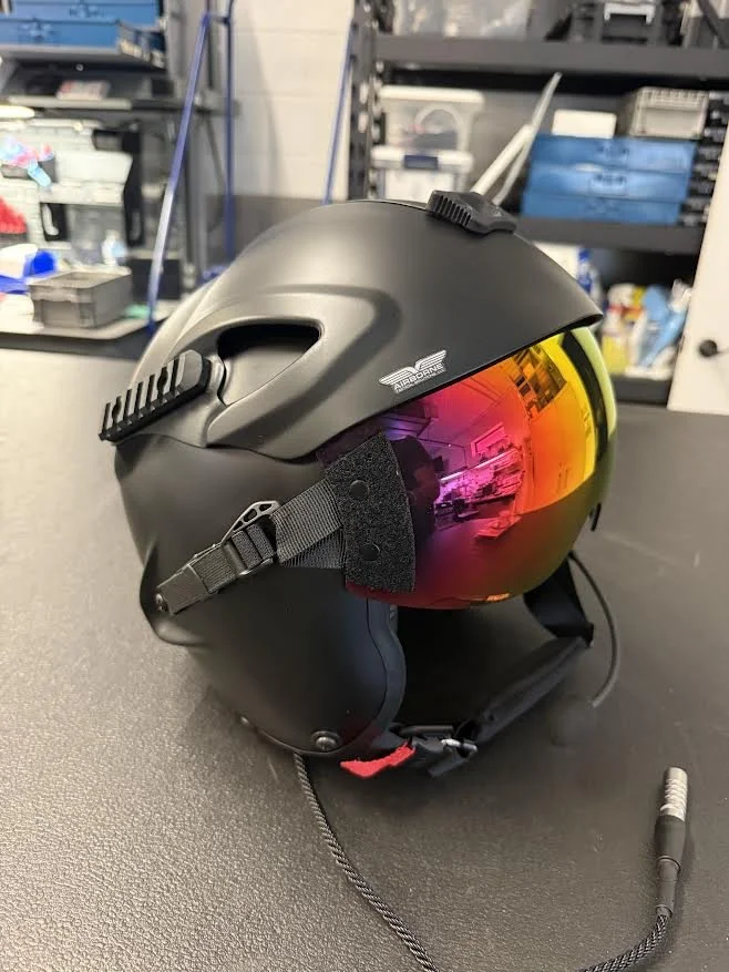 Black motorcycle helmet with a reflective orange visor and built-in communication equipment, placed on a black surface in a cluttered workshop or garage.