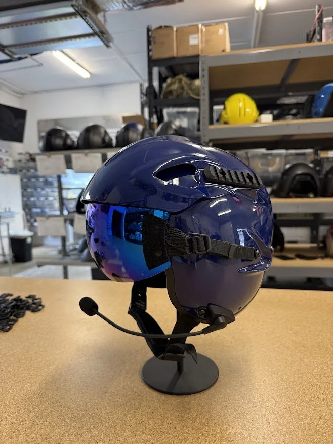 Blue bike helmet with built-in communication microphone on display in a storage room with helmets on shelves in the background.