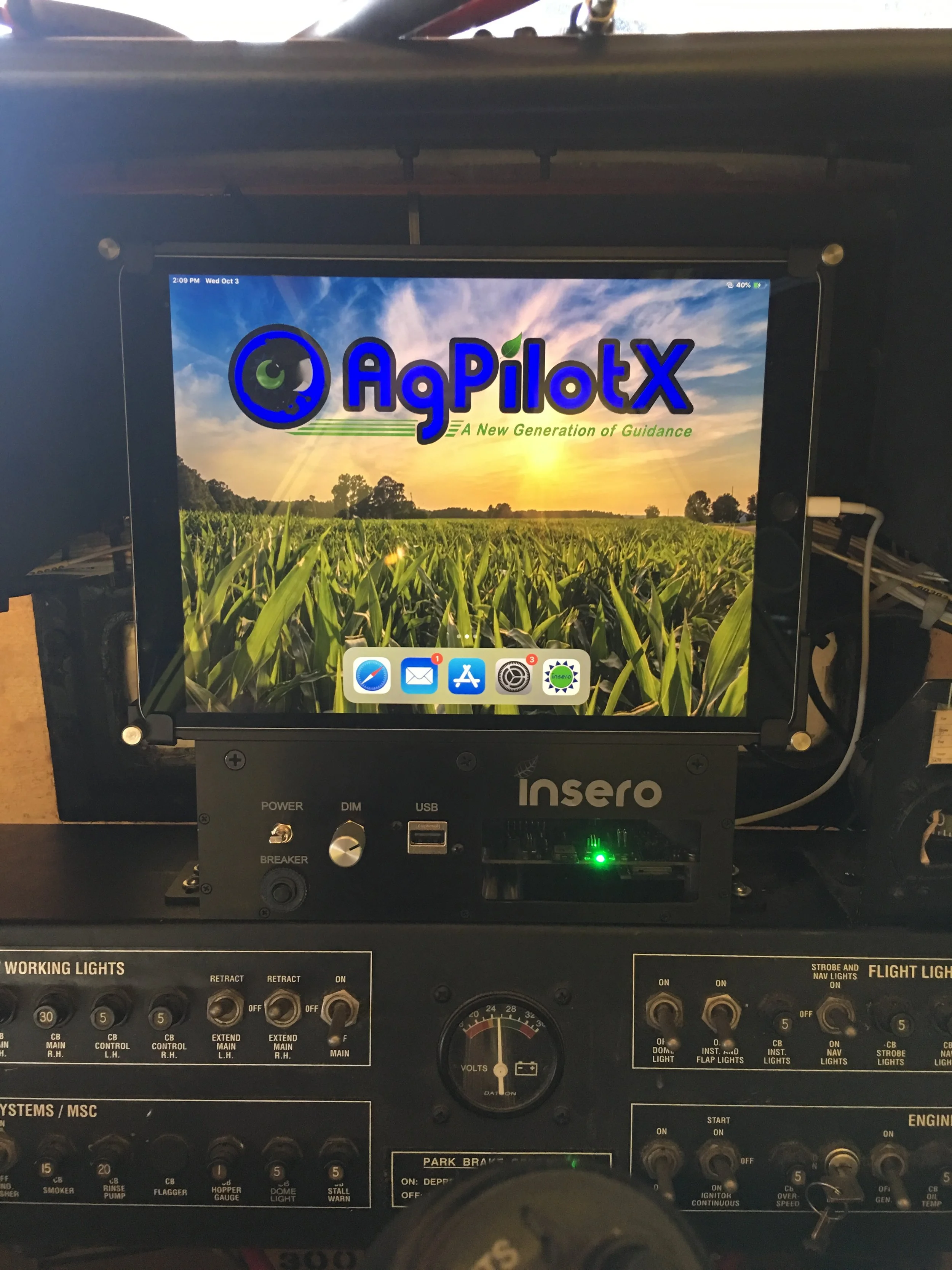 A flight simulator cockpit dashboard with a monitor displaying the logo 'AgPilotX' over a field landscape, with a sunrise or sunset in the background and various control switches and gauges in the foreground.