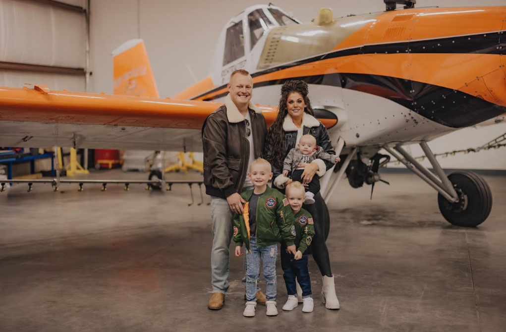 Family of five standing inside hangar with a large orange and black airplane in the background. The family includes a man, woman, and three young children, two of whom are wearing green jackets with NASA patches.