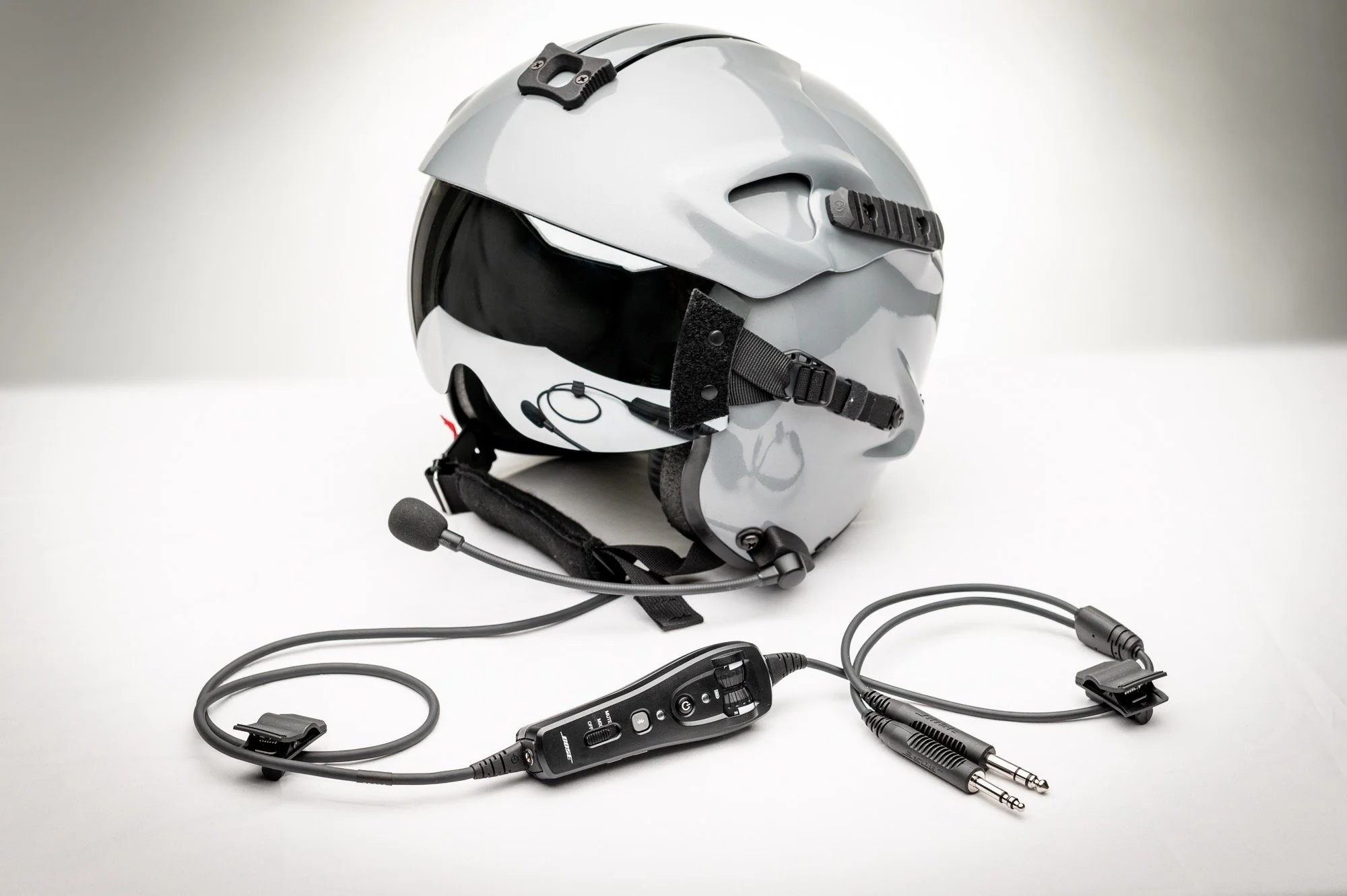A white motorcycle helmet with a black visor, attached communication device, and mounted microphone. In front of the helmet, there are various audio and communication cables, including a controller with multiple buttons and switchers, all arranged on a white surface.