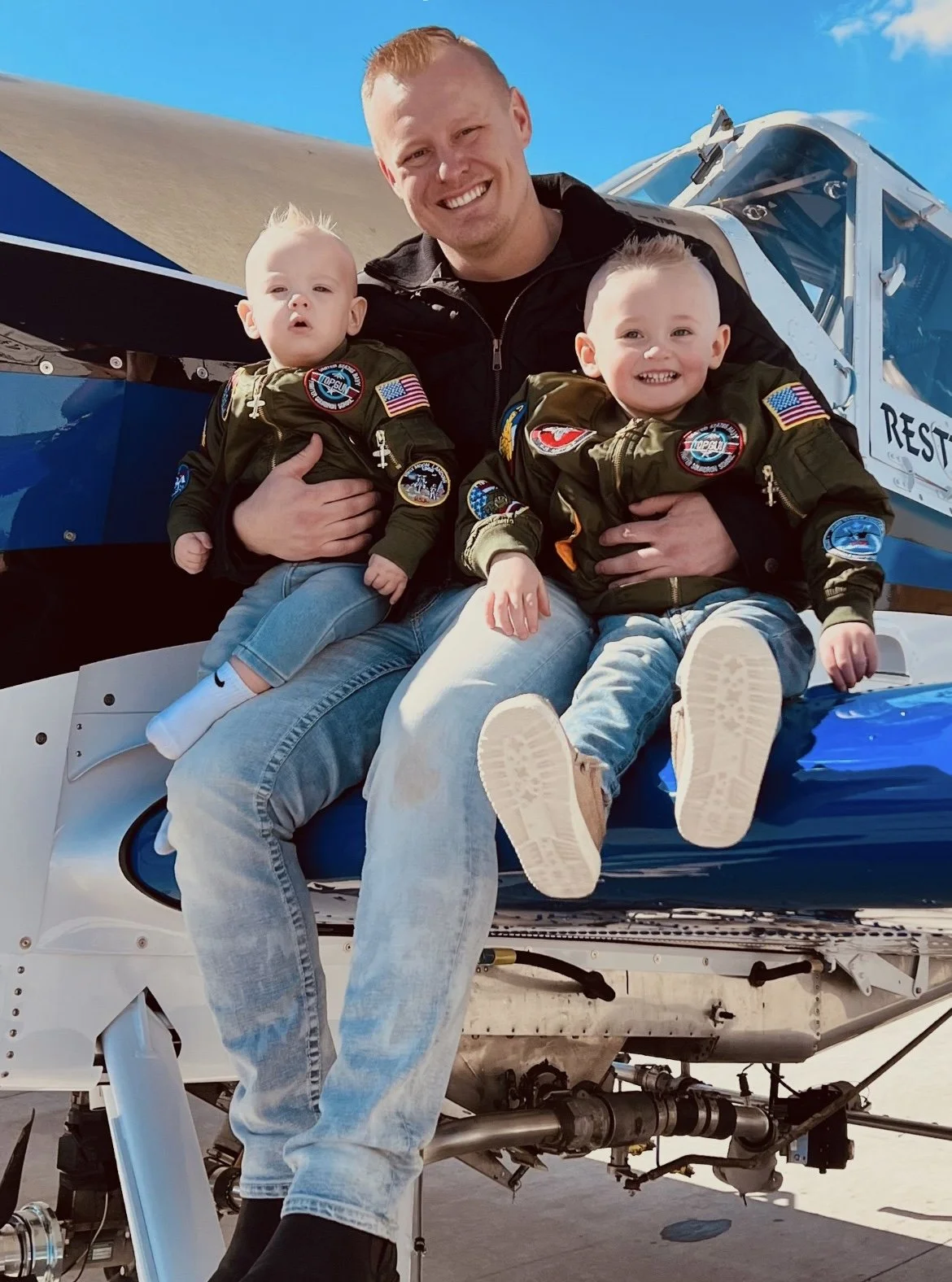 A man and two young children sitting on an aircraft, all wearing matching flight jackets with patches and smiling, during daytime outdoor setting.