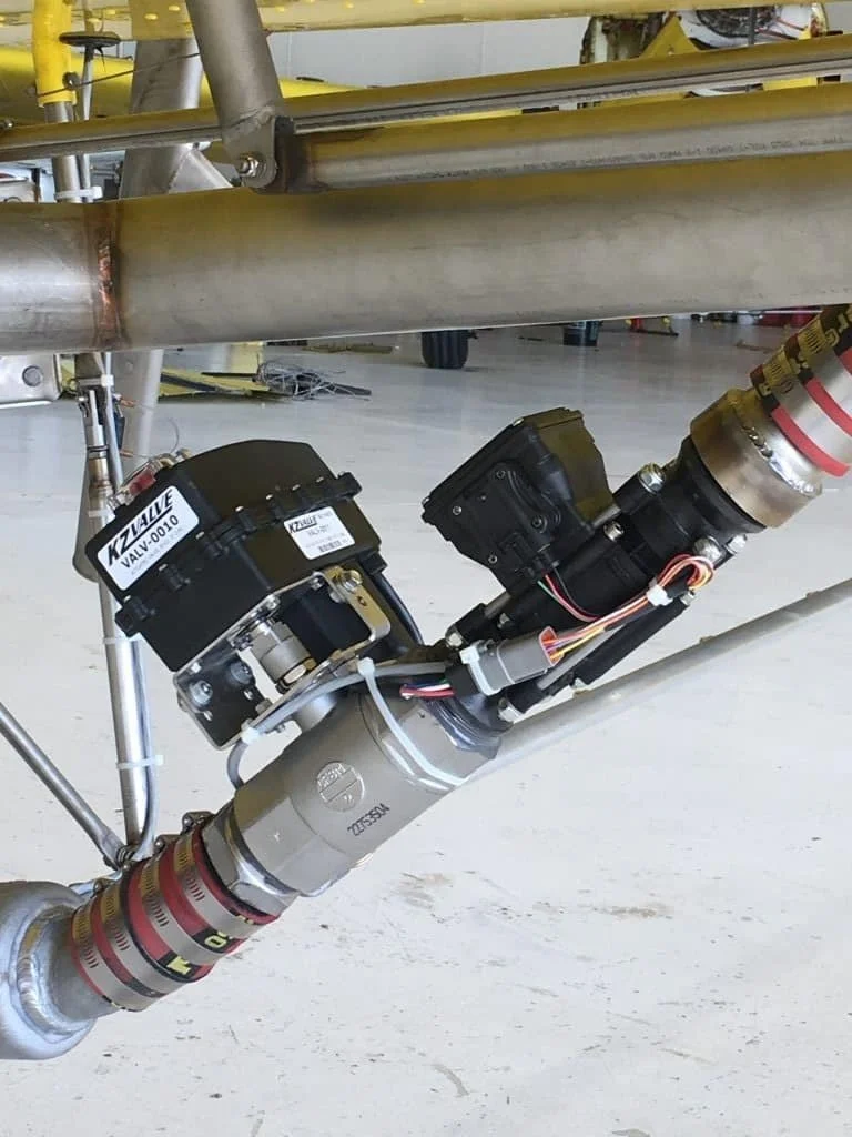 Close-up of a mechanical assembly with pipes, valves, and electronic components, including black valves with labels and wiring, on a metallic structure, likely part of industrial or aerospace equipment.