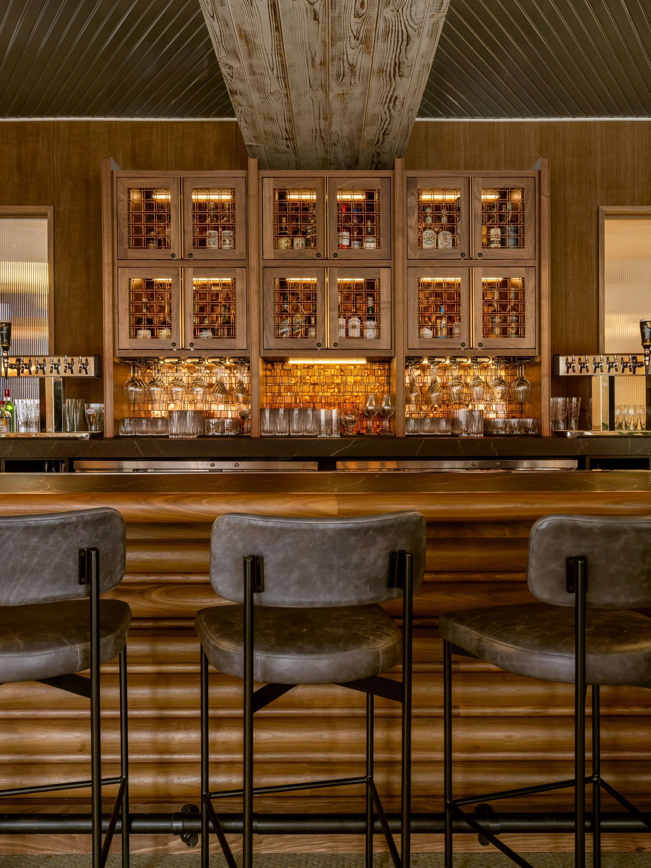 Bar area with wooden cabinets filled with bottles of alcohol and glasses hanging underneath, in a cozy, dimly lit setting.