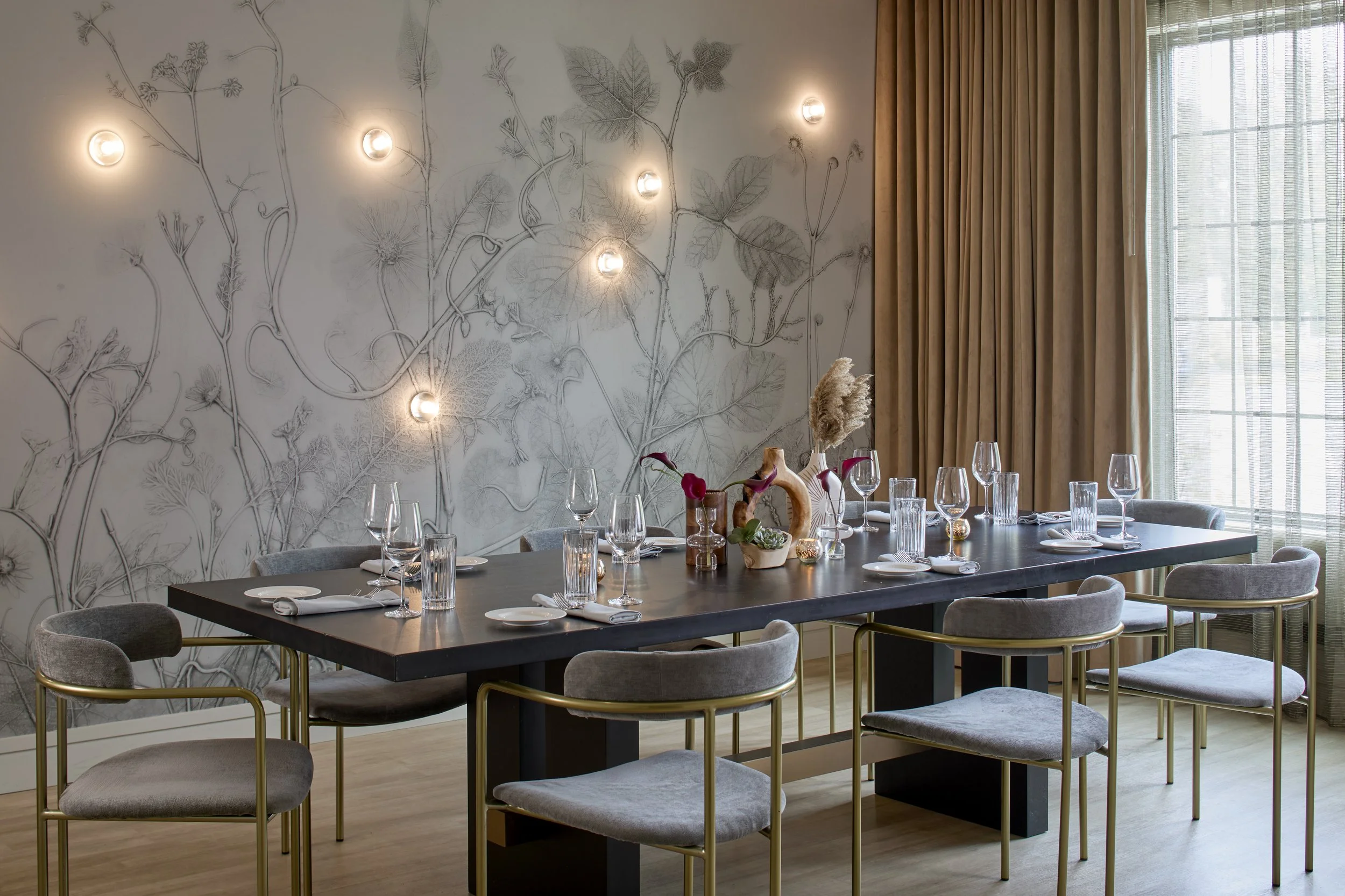 A dining room with a large black table set with wine glasses, plates, and napkins, surrounded by grey chairs with gold legs, and decorated with centerpieces including dried flowers. The wall has a large floral line drawing with wall-mounted lights, and there are beige and gold curtains covering windows.