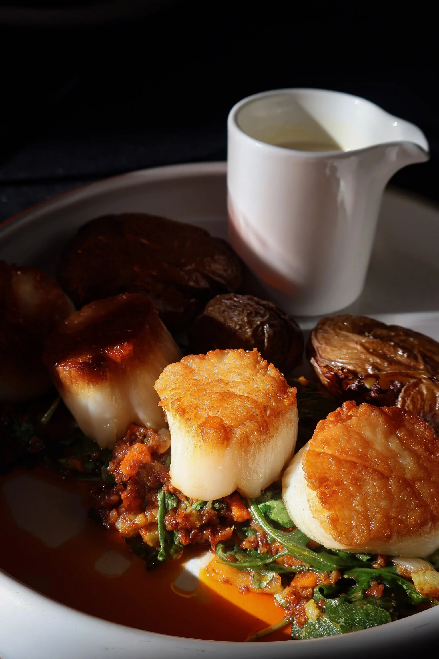 Roasted vegetables in a white bowl, including potatoes and root vegetables, with a small white pitcher of sauce in the background.