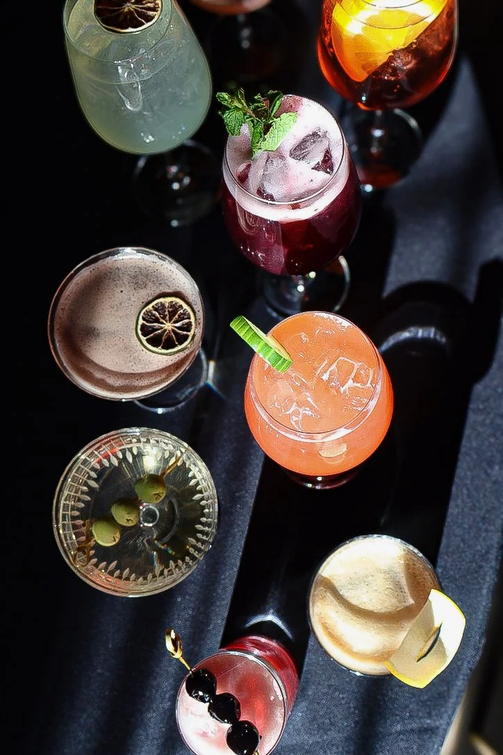 A variety of colorful cocktails and drinks on a black tray, garnished with fruit slices and herbs.