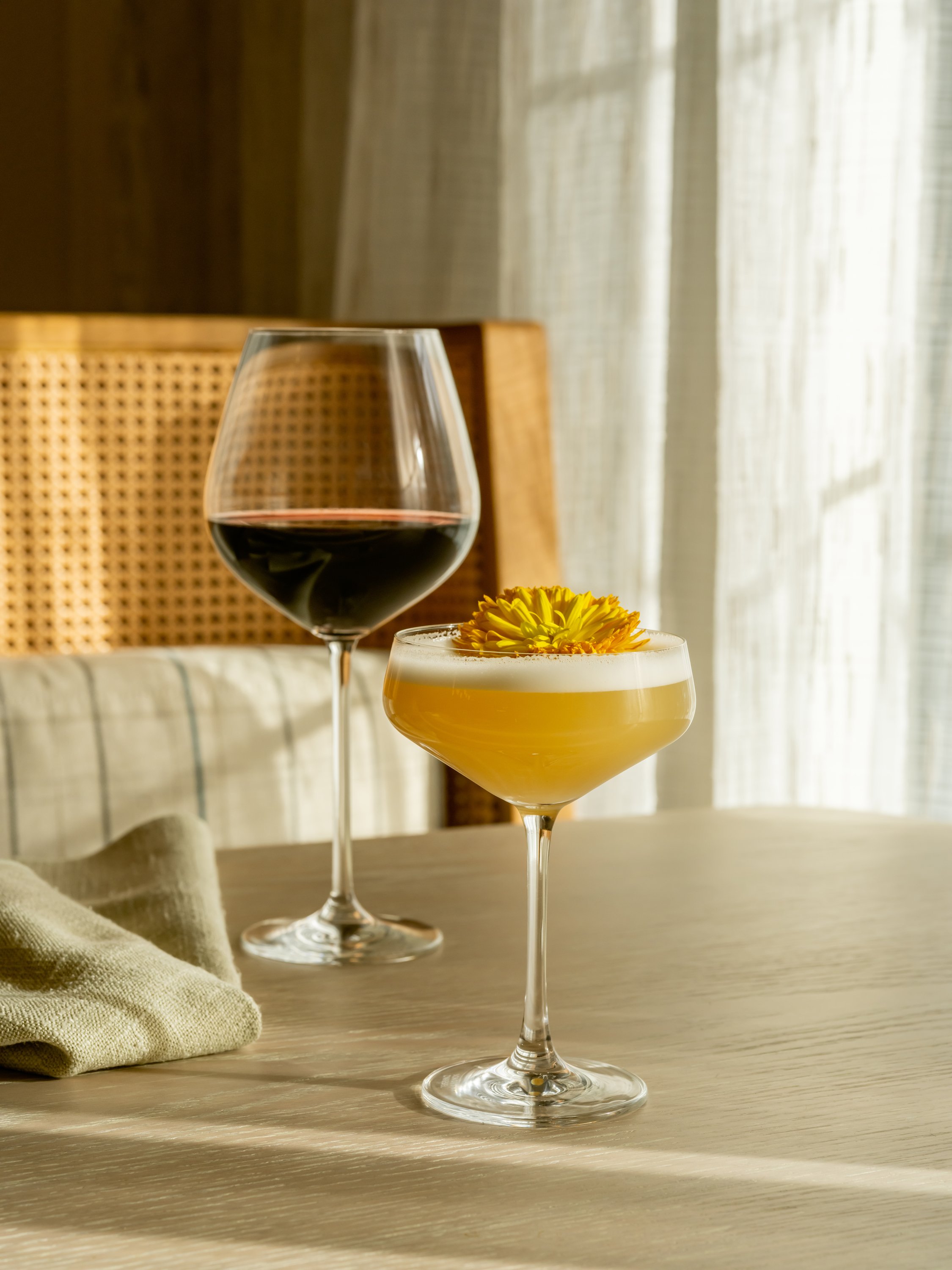 A glass of red wine and a cocktail with a yellow flower garnish on a wooden table, sunlight streaming through a window with curtains in the background.