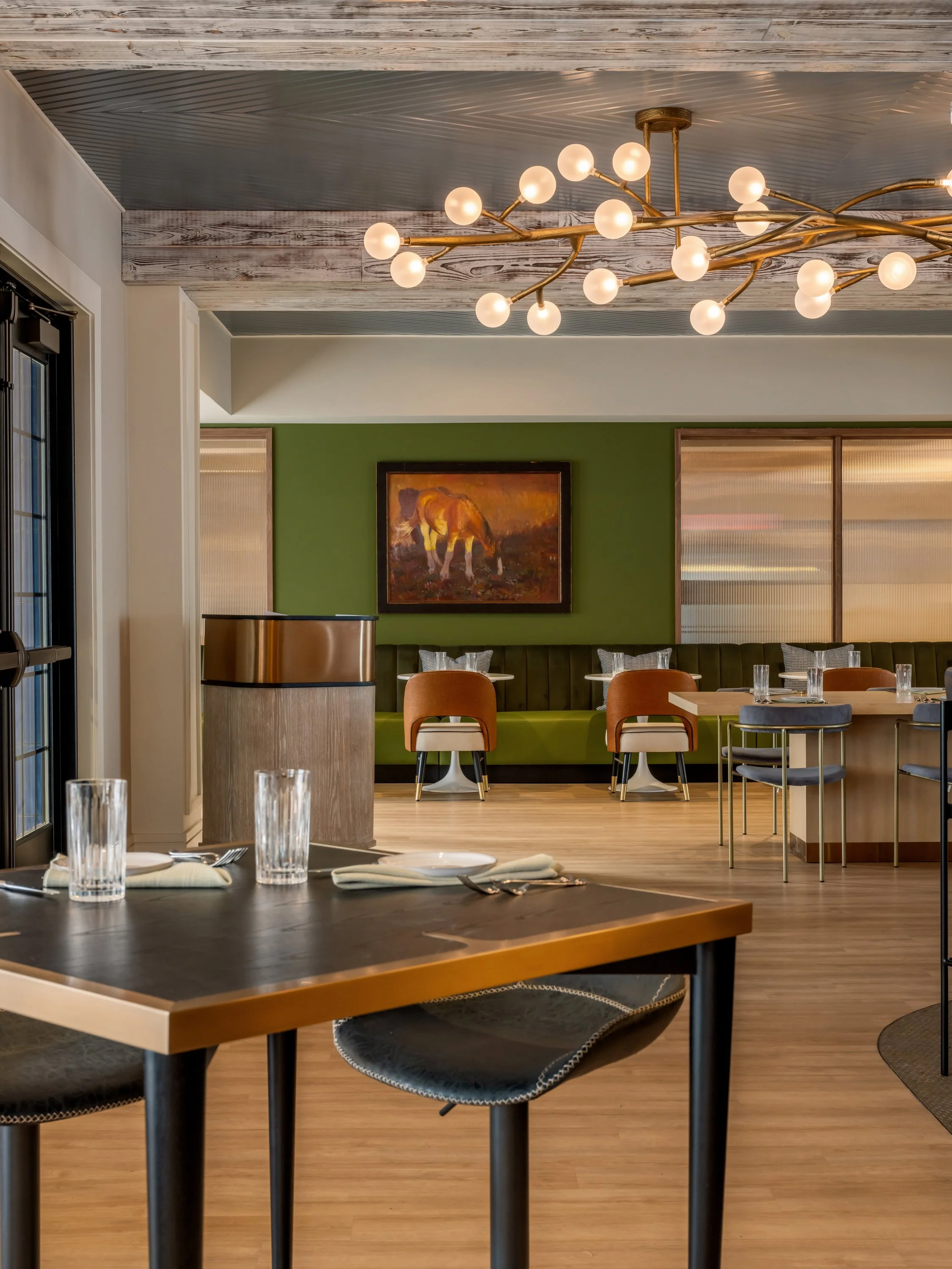 Interior view of a restaurant with a green wall, a painting of a horse, tables set with glasses and silverware, and a modern chandelier with multiple round bulbs.