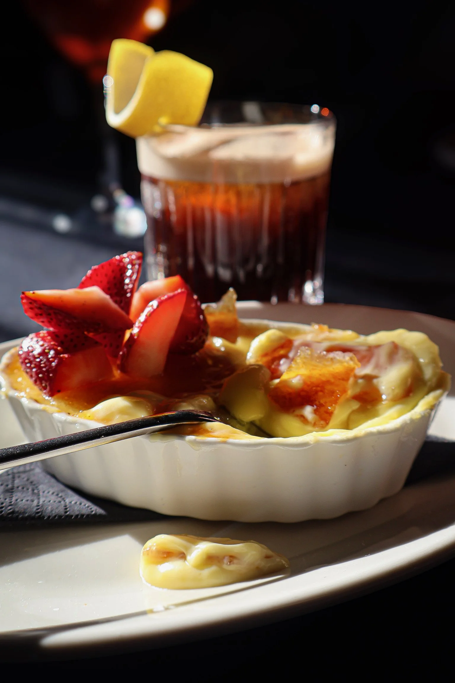 A baked dish with melted cheese, slices of strawberries, and sliced fruit on top, served in a white dish with a spoon. There is a glass of dark beverage with lemon wedge garnish in the background.