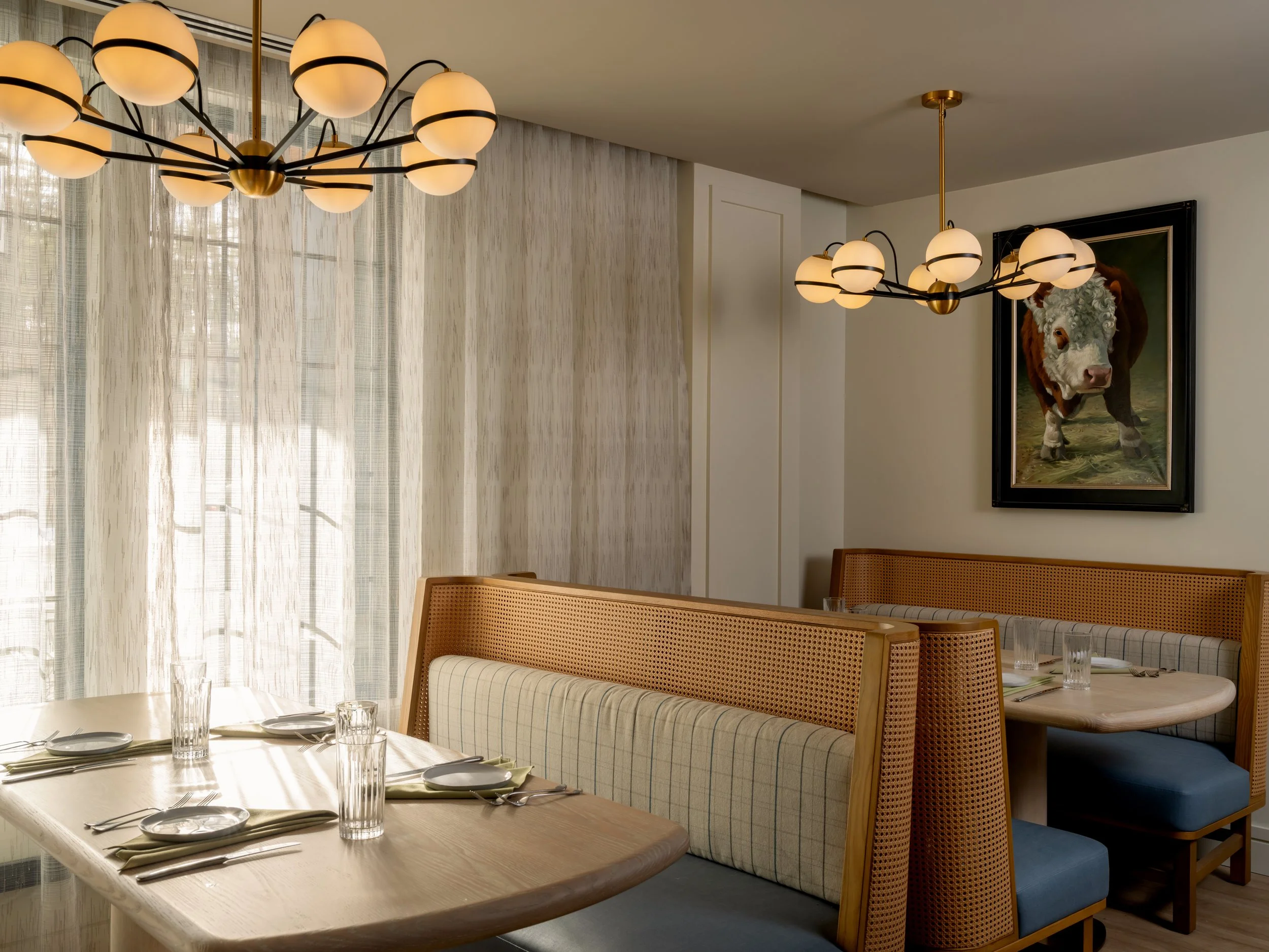Dining area with a table set with glasses, plates, and napkins. There are upholstered chairs and a booth with a checked pattern. Two modern chandeliers hang from the ceiling, and sheer curtains cover the windows, allowing light to filter through. A f